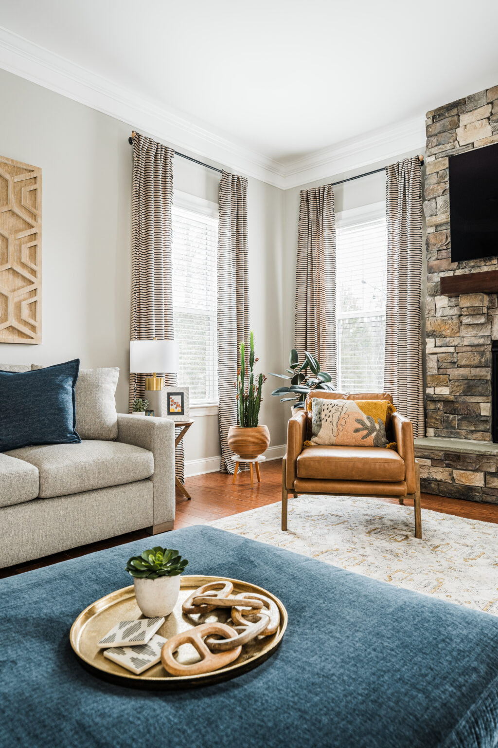Living room with a stone fireplace, a tan leather accent chair, and a navy ottoman, by Hayden Home Studio in Weddington, NC